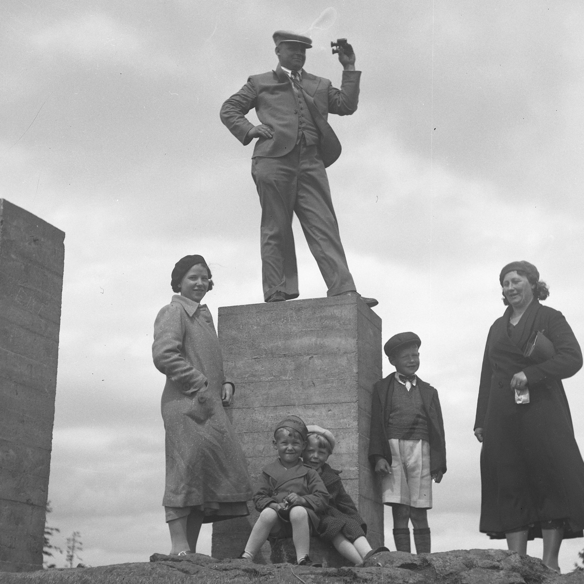 En man står på en hög stenplatta med sin kikare. Nedanför står hans familj.