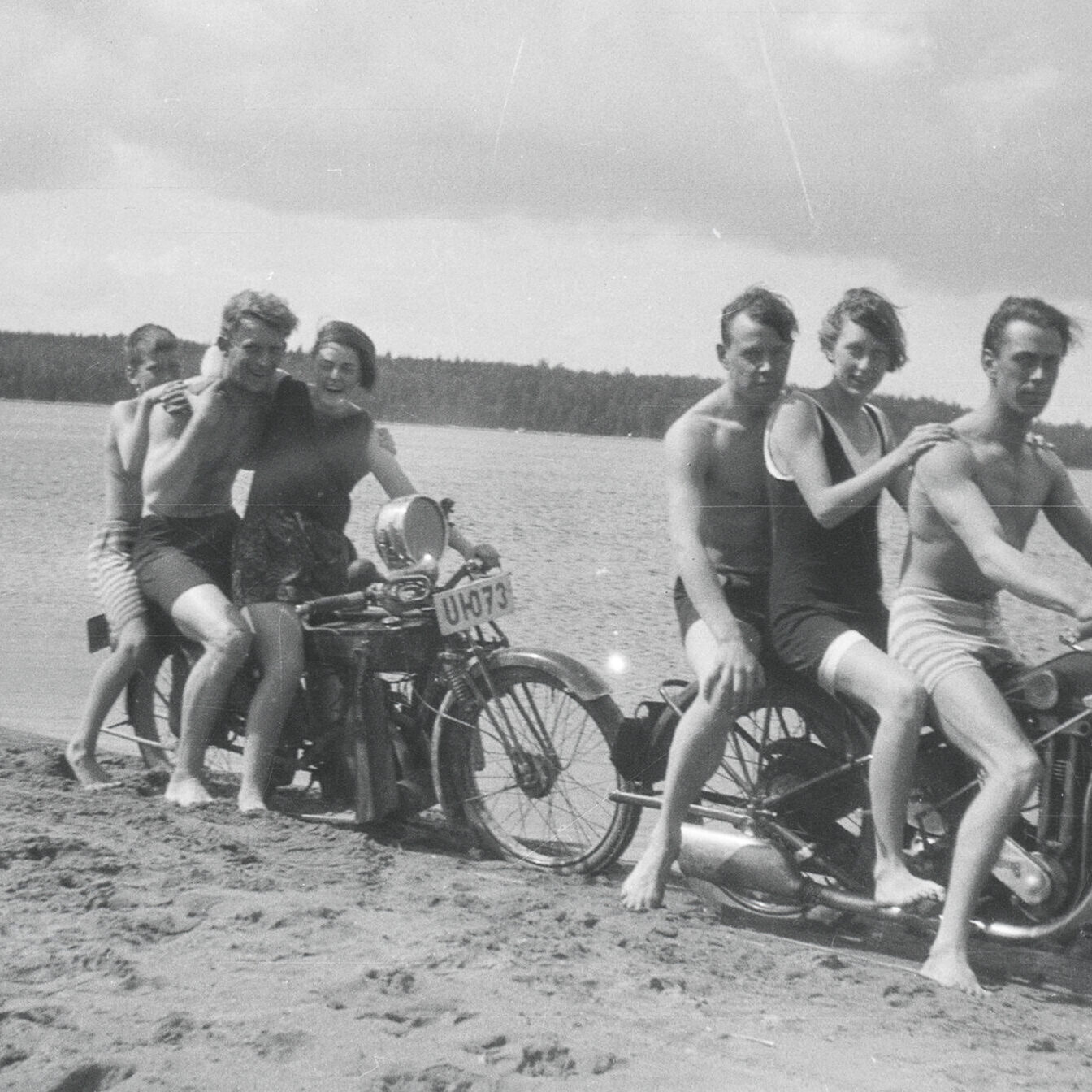 Sex badklädda personer på två motorcyklar vid en sjöstrand.